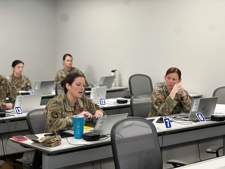 Woman in Air Force uniform appears to be asking a question while gesturing towards her computer in a classroom with other military members