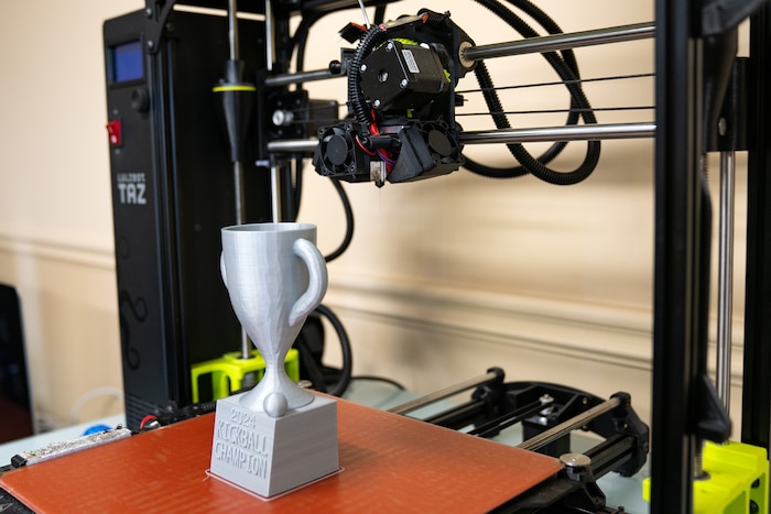 A grey plastic trophy sits under the arm of a 3D printer