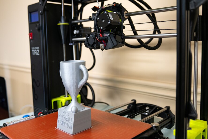 A grey plastic trophy sits under the arm of a 3D printer