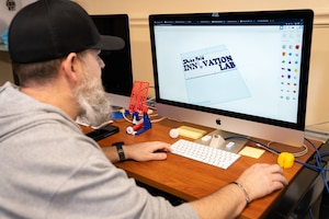 A bearded man with a ballcap and a sweatshirt sits in front of a computer operating a 3D design program. In the window he is viewing a sign for the innovation lab he created featuring a lightbulb as the first "O' in innovation. He's surrounded by 3D printed trinkets on a wood desk top.