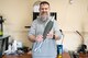 Bearded man in sweatshirt holds up a faux plastic armament with a dull base and shiny metallic features at both ends. He's surrounded by scattered cords, miscellaneous items, and technology on a desk.
