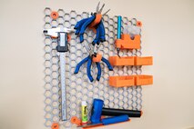 Colorful blue and orange tools hang from a mesh hexagonal structure from a wall.