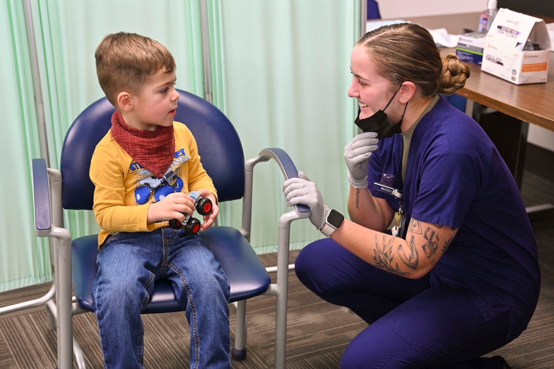 A child sits in a chair talking to an airman in scrubs crouche next to him.