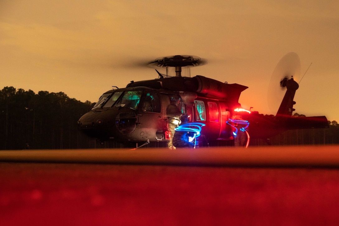 A Black Hawk helicopter sits on a flight line bathed in red and blue lights with rotors going.