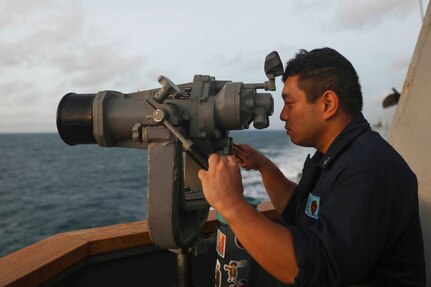 A service member in uniform looks through binoculars aboard a Navy vessel.