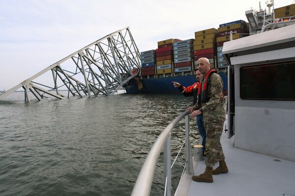 Two people on white boat with grey metal railing overlook a collapsed bridge while a blue cargo ship with red, yellow and green containers in background