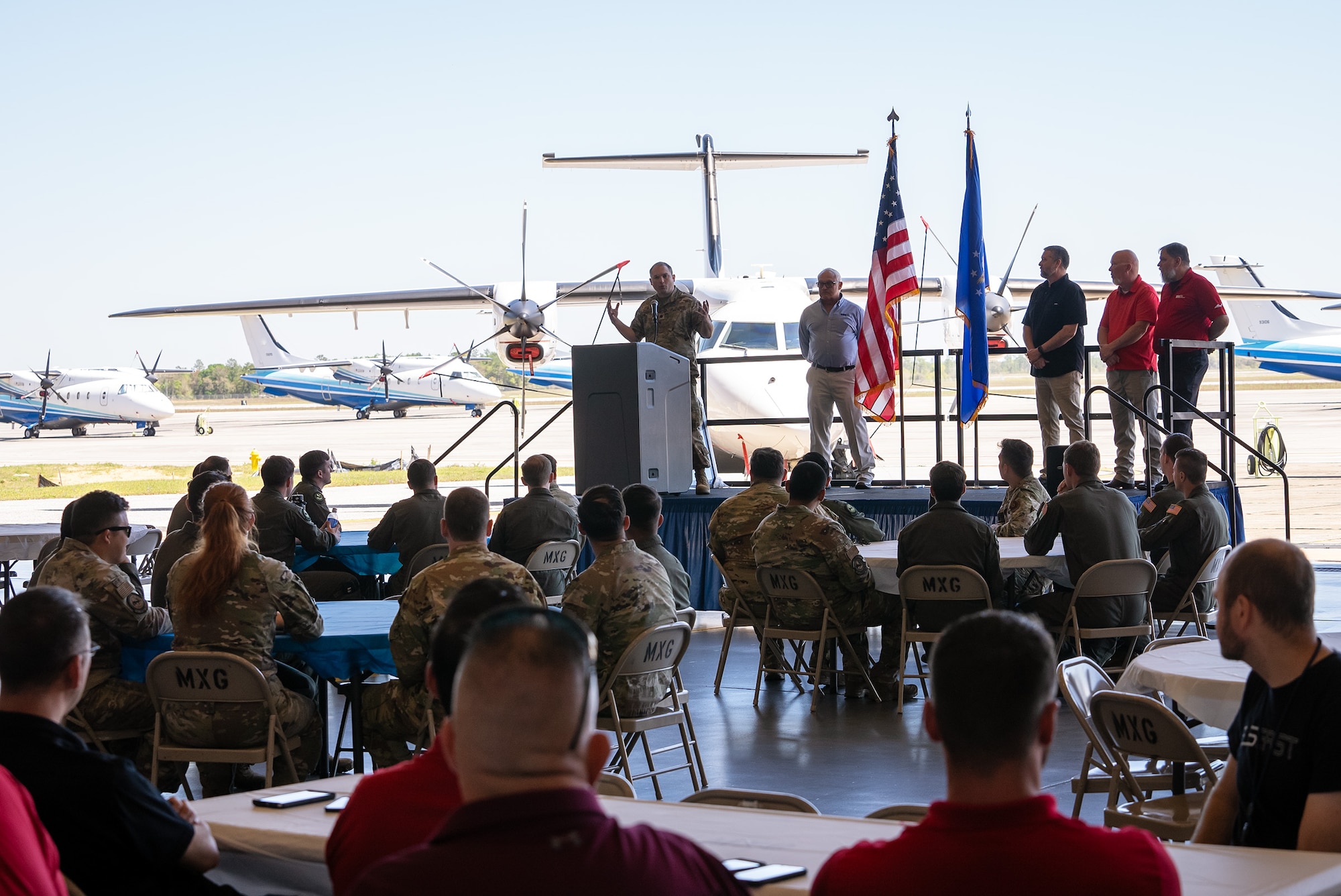 A group of people stand on a stage give a presentation in front of multiple airframes to a gathering of civilians and military