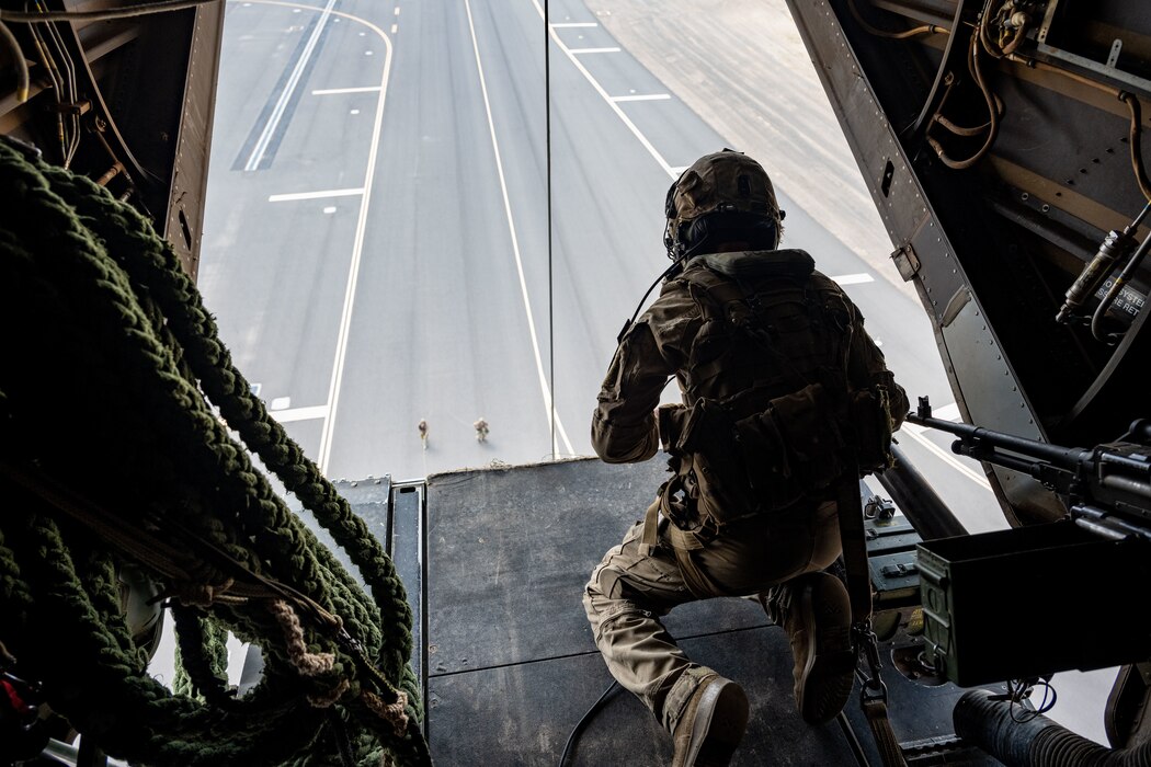 A U.S. Marine in aircrew uniform looks out the open cargo ramp of an MV-22 Osprey in flight.