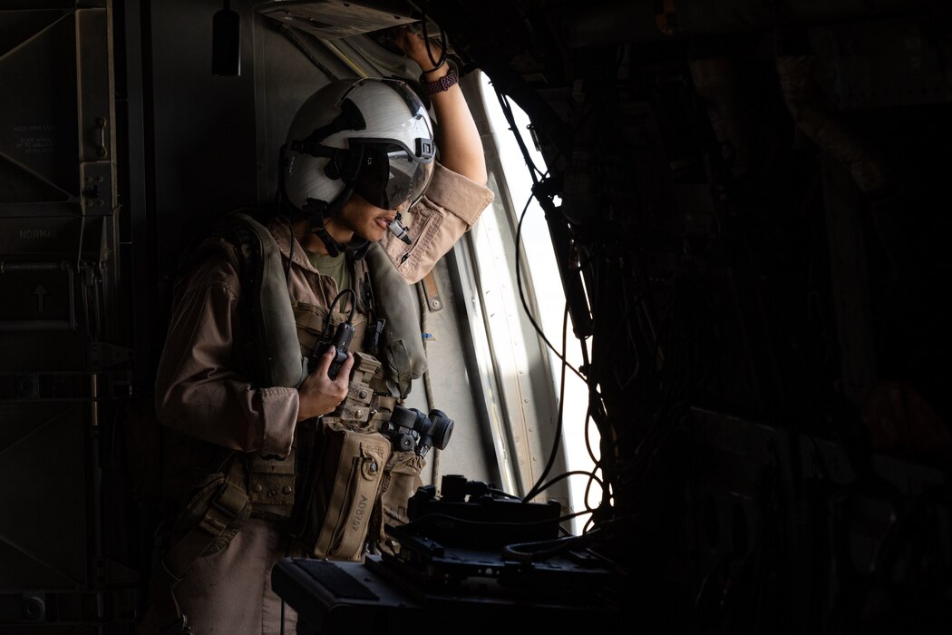 An Airman wearing flight gear with a helmet and visor looks out an aircraft window.