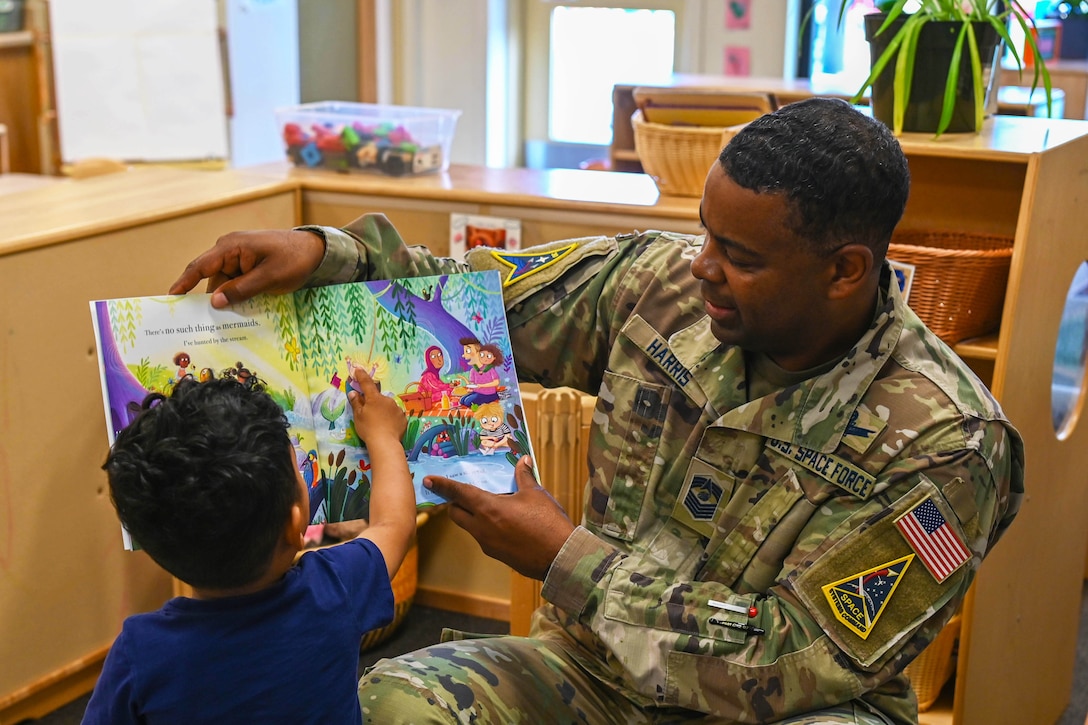 A uniformed service member holds a book and reads to a child whois pointing at an illustration in the book.