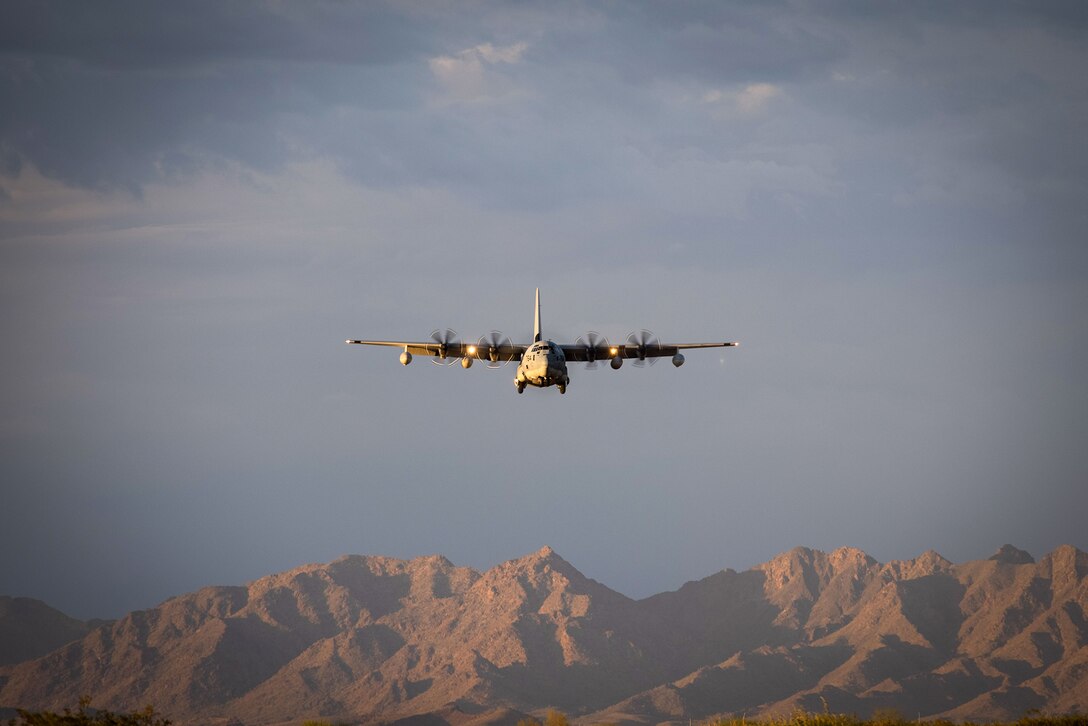 A military aircraft flies toward the camera during daylight with mountains in the background.
