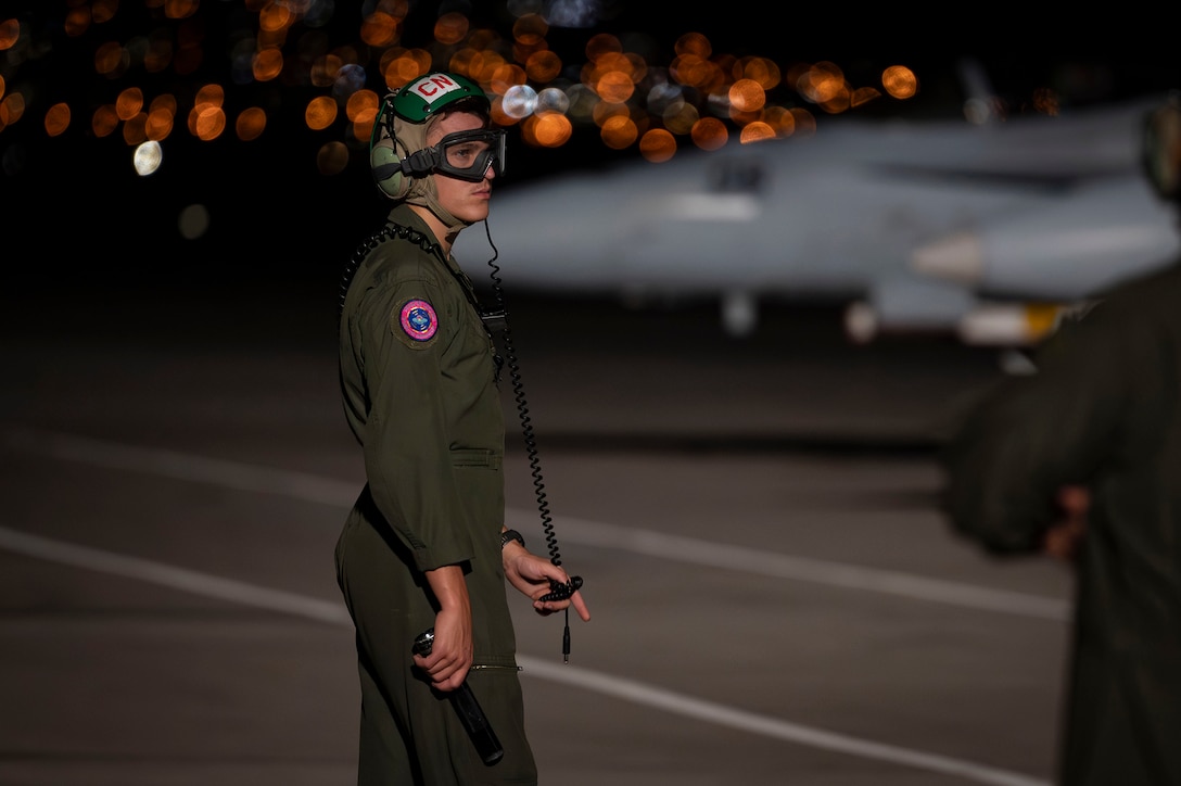 A service member in a flight suit and protective headgear and eyewear stands on a tarmac at night with a military aircraft parked in the background.