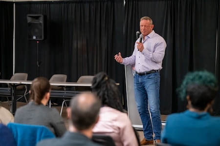 A man wearing jeans and a light blue dress shirt gives a presentation in front of an audience.