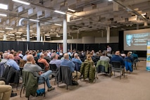 An audience sits and watches a presentation in a large room.