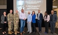 A group of nine people stand on a small stage to pose for a photo. Behind them is a large projection screen reading "A Male Perspective with Keynote Speaker Clark Federicks.