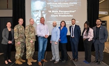 A group of nine people stand on a small stage to pose for a photo. Behind them is a large projection screen reading "A Male Perspective with Keynote Speaker Clark Federicks.