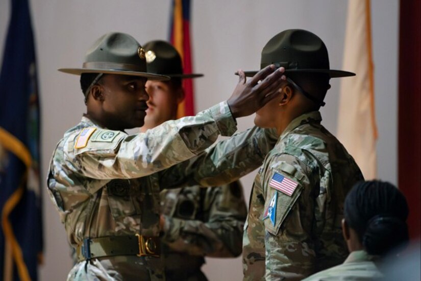 A uniformed service member adjusts the drill sergeant hat of another during a graduation ceremony.