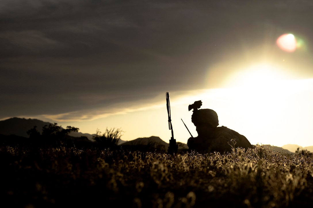 Seen in silhouette, a Marine in tactical gear lays in grass in a mountainous area while speaking on a radio as light shines from the right.