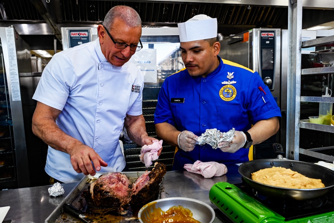 A sailor in a culinary uniform speaks to a chef while preparing food in a kitchen.