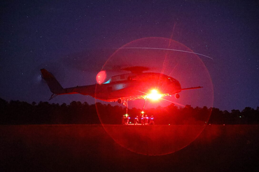 A circle of red light reflects over the scene as Marines hold ropes connected to a helicopter hovering over a field under a starry sky.