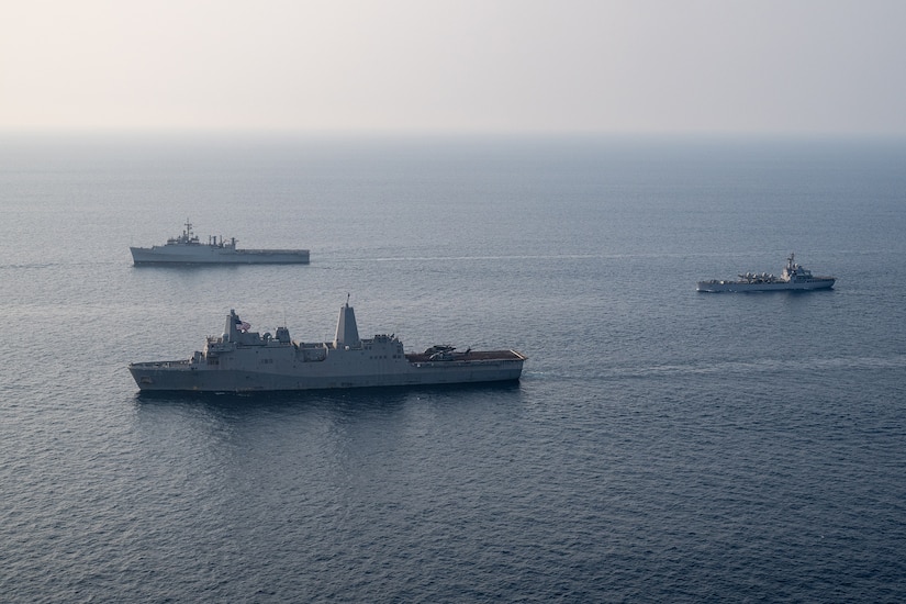 Three gray Navy ships steaming in formation across a relatively calm ocean.