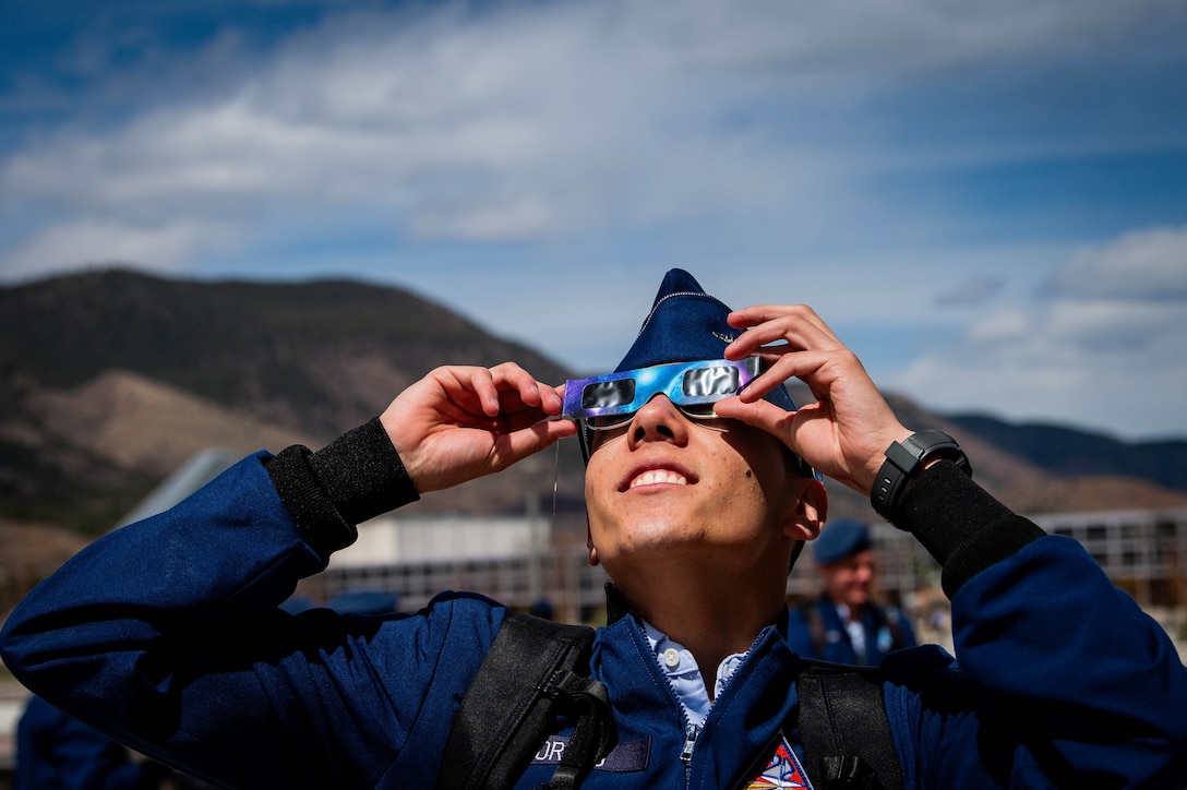 An Air Force cadet holds up solar glasses over their sunglasses while looking up toward the sky with cloudy skies and mountains in the backdrop.