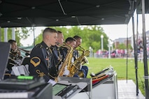 Several Army soldiers dressed in dark ceremonial uniforms are performing on musical instruments under a dark tent at an outdoor event. There is a large green lawn in the background.