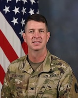 White man with ashort dark brown hark in Army green camouflage inform posing for a picture in front of the US Flag.