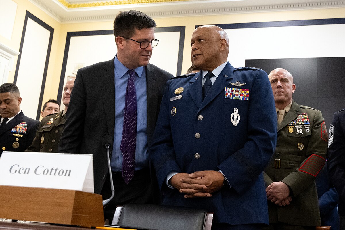 A person in a suit stands next to another person in a military uniform.