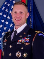 White male with short blond hair dressed in dark Army service uniform (dark jacket and tie, white shirt, several ribbons and badges, and a gold-colored rank on shoulders) smiling while posing for a picture in front of the US Flag and another dark blue flag.