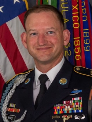 White man with short light brown hair wearing Army service uniform (dark jacket, dark tie, white shirt, multicolored ribbons and badges) smiling while posing for a picture in front of the US flag and some multicolored streamers with illegible text on them.