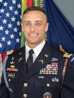 White man with shorter dark brown hair wearring the Army service dress uniform (dark jacket and tie, white shirt, many multicolored ribbons and metal badges) is smiling while posing for a picture in front of the U.S. flag and a dark blue flag with multicolored streamers hanging from the top of the staff.