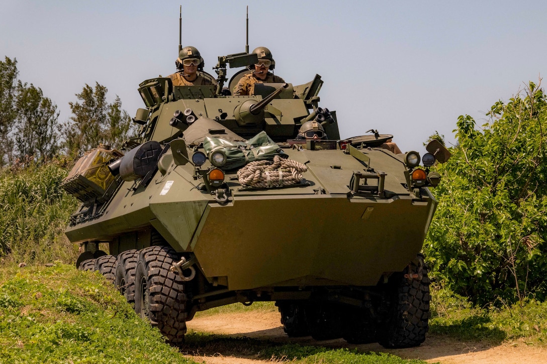 Two Marines ride in an armored vehicle on a dirt road through the woods.