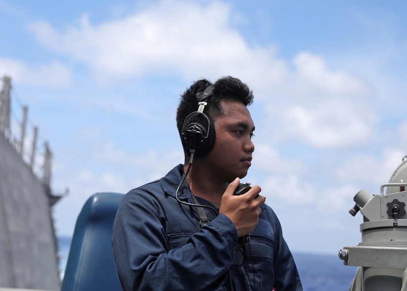 A sailor holds a radio and wears headphones with a ship and water in the background.