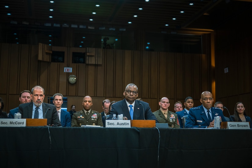 Three men, two in business suits and one in a dress military coat, sit at a long table looking forward. Two service members in dress uniforms sit behind them, as do a handful of civilians.