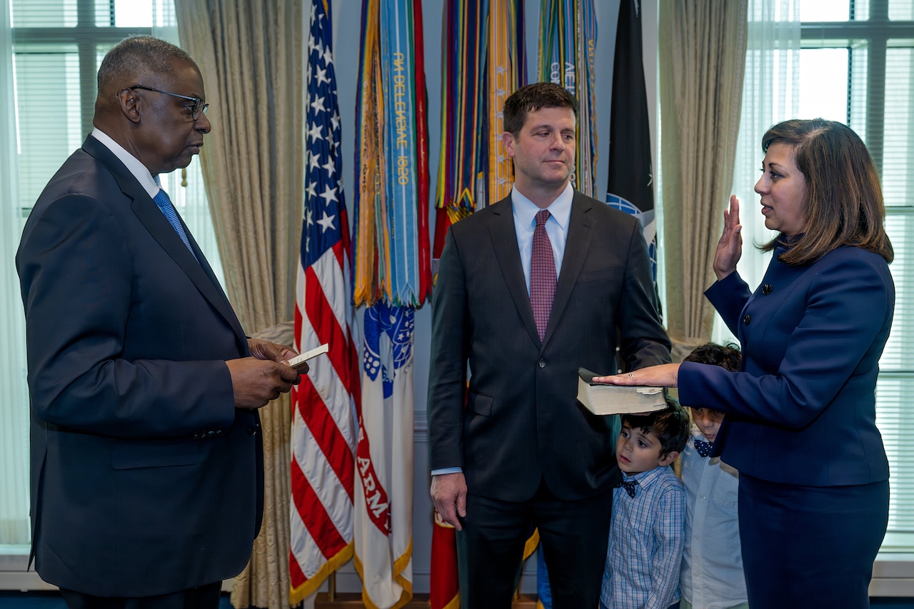 Secretary of Defense Lloyd J. Austin III faces another civilian who stands with right hand raised and left hand on a book a third person holds.