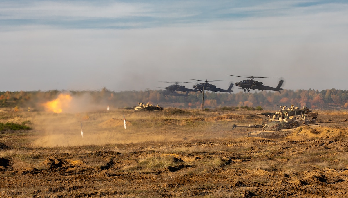 Three helicopters fly over a large field where military tanks fire ammunition.