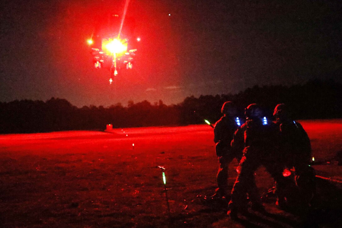 Three Marines on the ground use a flare to guide a helicopter in the air with its lights on at night. The scene is bathed in a red light.