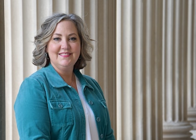 A blond woman in a teal jacket looks at the camera and smiles while she stands in front of marble pillars, she is outside.