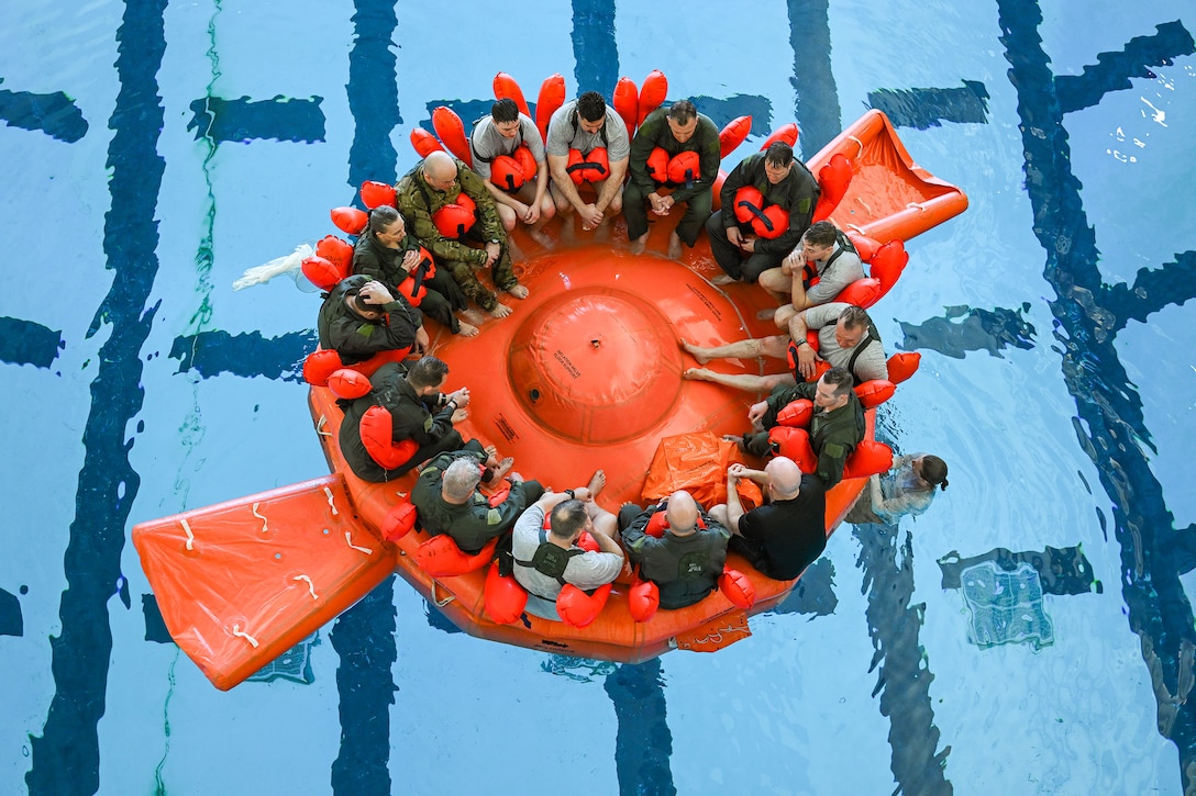 A group of reservists are seated in a 20-person life raft in a pool during training as one person swims outside the raft.