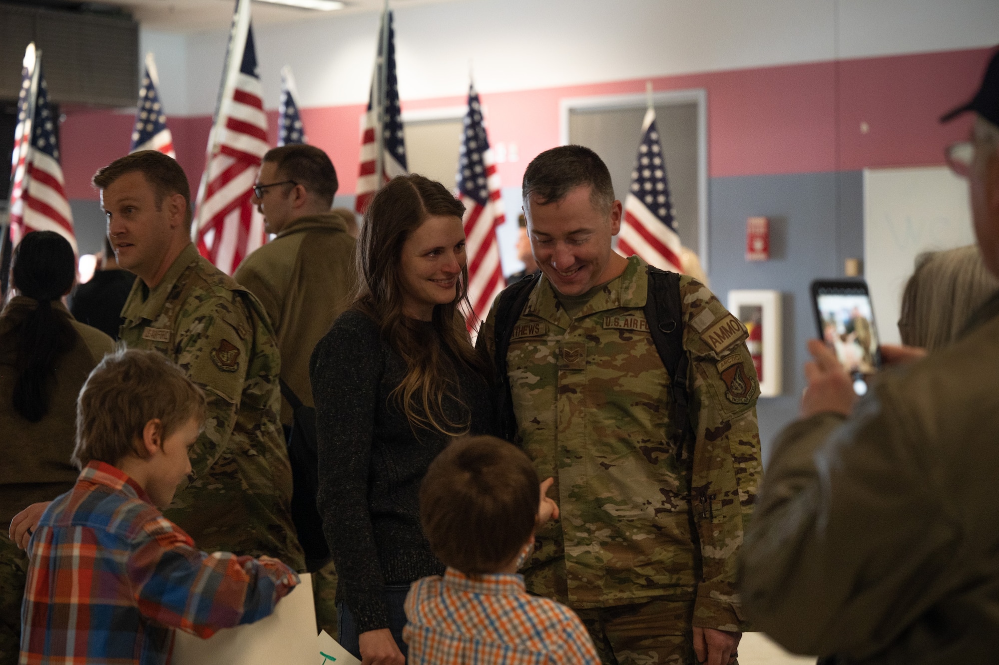 A father smiles down at his son as his son points towards him. His wife stands beside him smiling. The family is surrounded by several other U.S. Air Force members reuniting with their friends and family.
