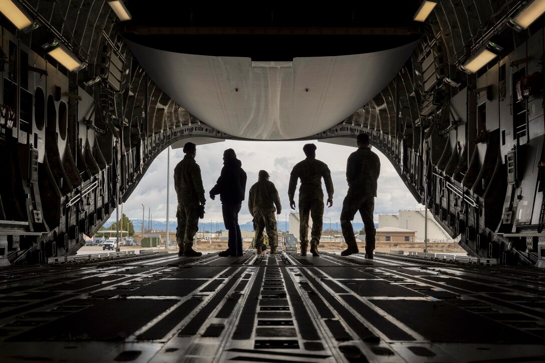 Four airmen stand in the open doorway of an aircraft as seen in silhouette with buildings and mountains in the background.