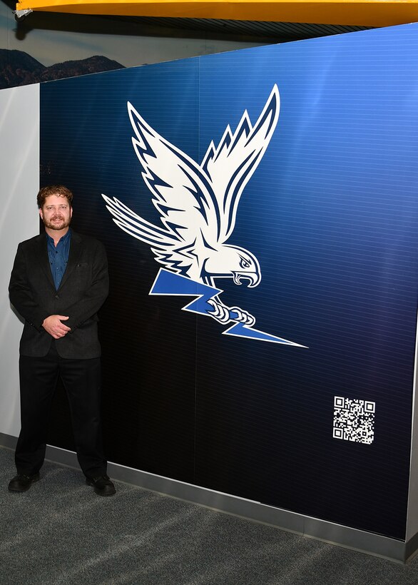 A man in a dark suite stands next to a graphic of an eagle representing the US Air Force. The graphic is on a wall inside a building.