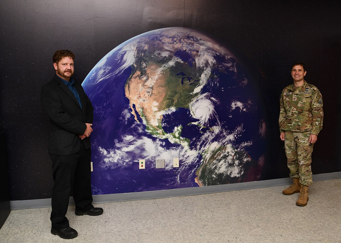 A man in a dark suit stands on the left side of a wall print of the earth and a man in a military camo uniform stands on the right side of the print.
