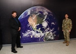 A man in a dark suit stands on the left side of a wall print of the earth and a man in a military camo uniform stands on the right side of the print.