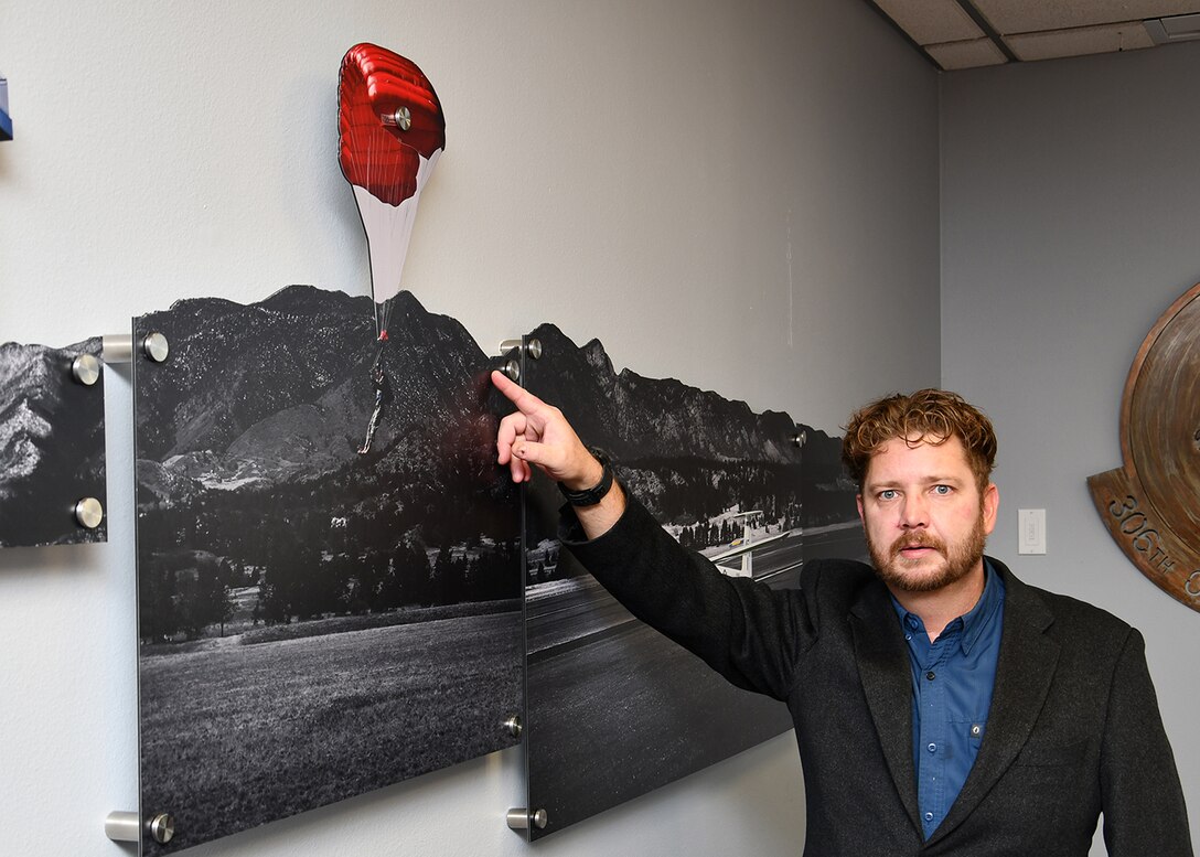 A man with a beard and dark jacket points at wall art. the are is a black and white photo print where the sky has been cut out. The print is not flush to the wall but mounted on to pegs that hold it a few inches from the wall giving it more depth.