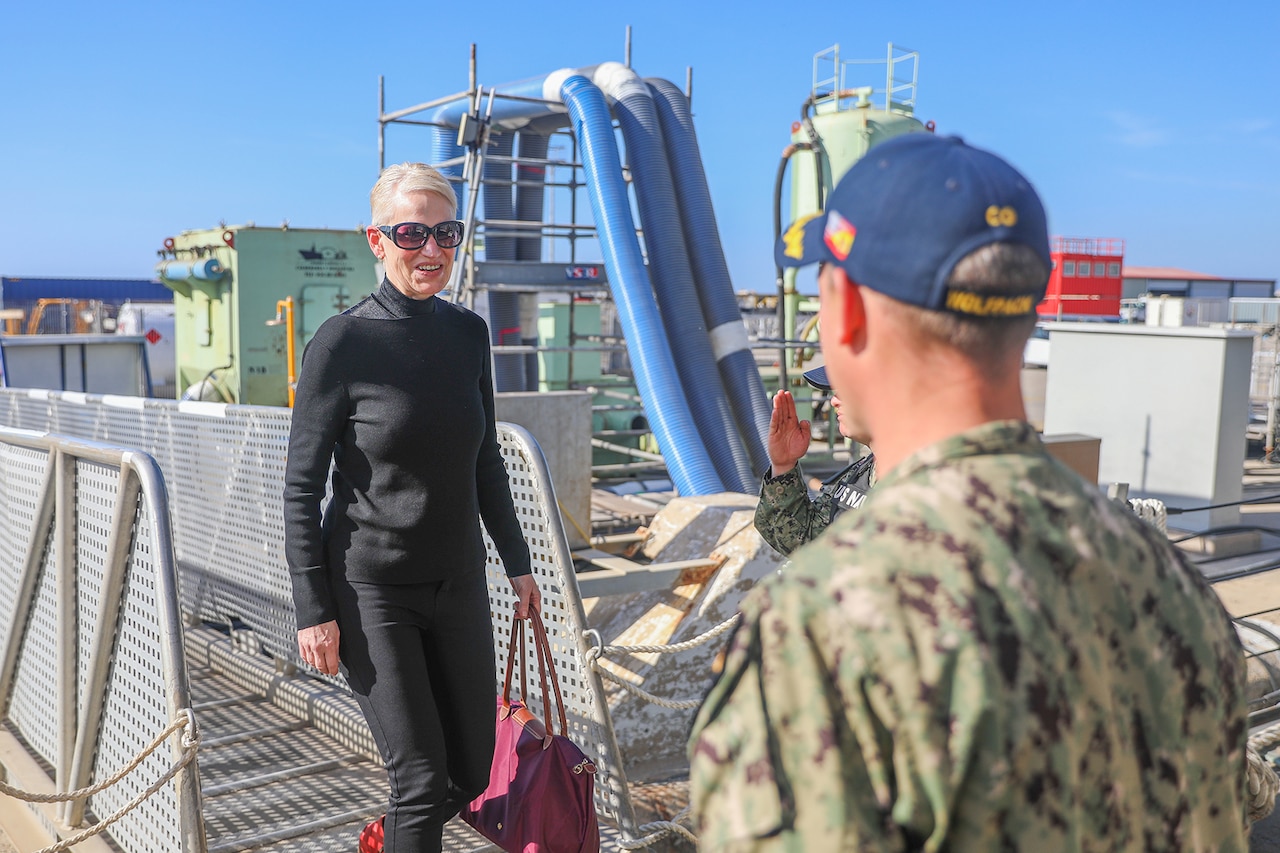 A person in civilian clothing and sunglasses smiles while approaching a uniformed service member on a ship.