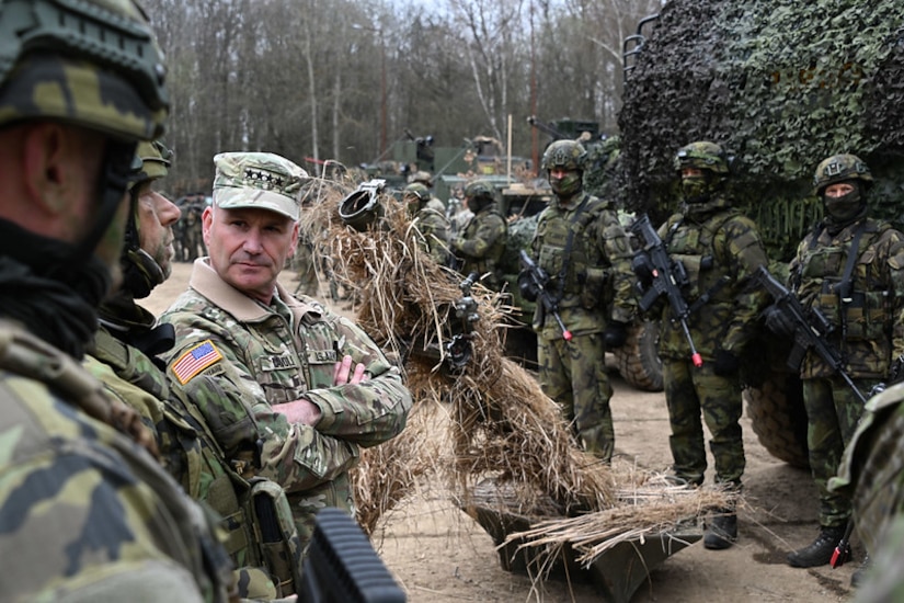 A man in uniform with a hat that has four stars is surrounded by service members in uniform holding weapons. Military equipment is camouflaged in hay and tarps.