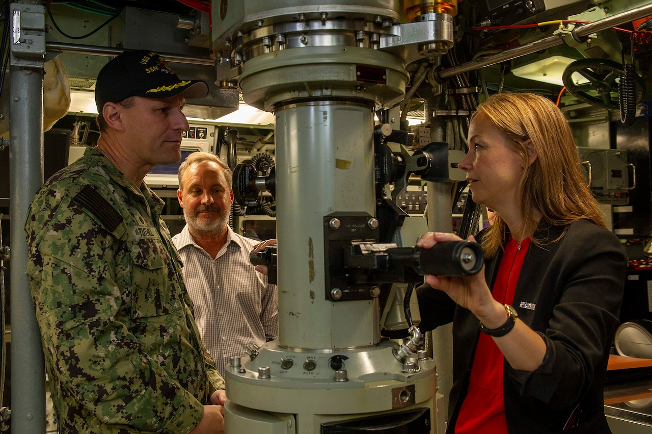 A person in civilian attire speaks with a sailor in uniform next to a periscope aboard a submarine.