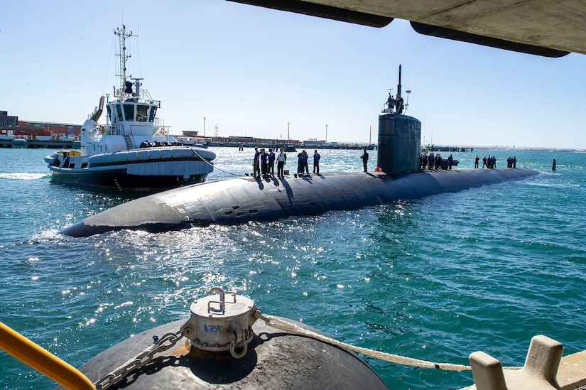 Sailors stand aboard a submarine while afloat near a pier.